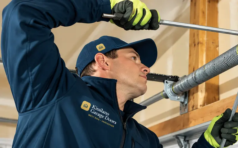 Kitsilano Garage Doors technician in a navy uniform adjusting a torsion spring above a residential garage door
